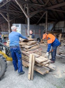Ein Radlader hat mit seiner Gabel einen großen Bretterstapel geladen. Drei Männer arbeiten daran: 2 Männer verladen das Holz, 1 Mann sägt die Bretter mit einer Motorsäge kleiner