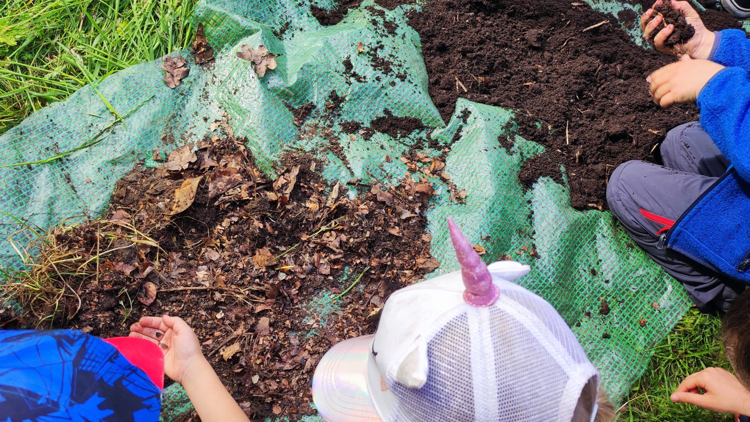 Kinder sind über einen Haufen feinen und einen Haufen groben Kompost gebeugt und untersuchen ihn. Ein Kind hat eine Assel in der Hand, ein anderes die feine Erde