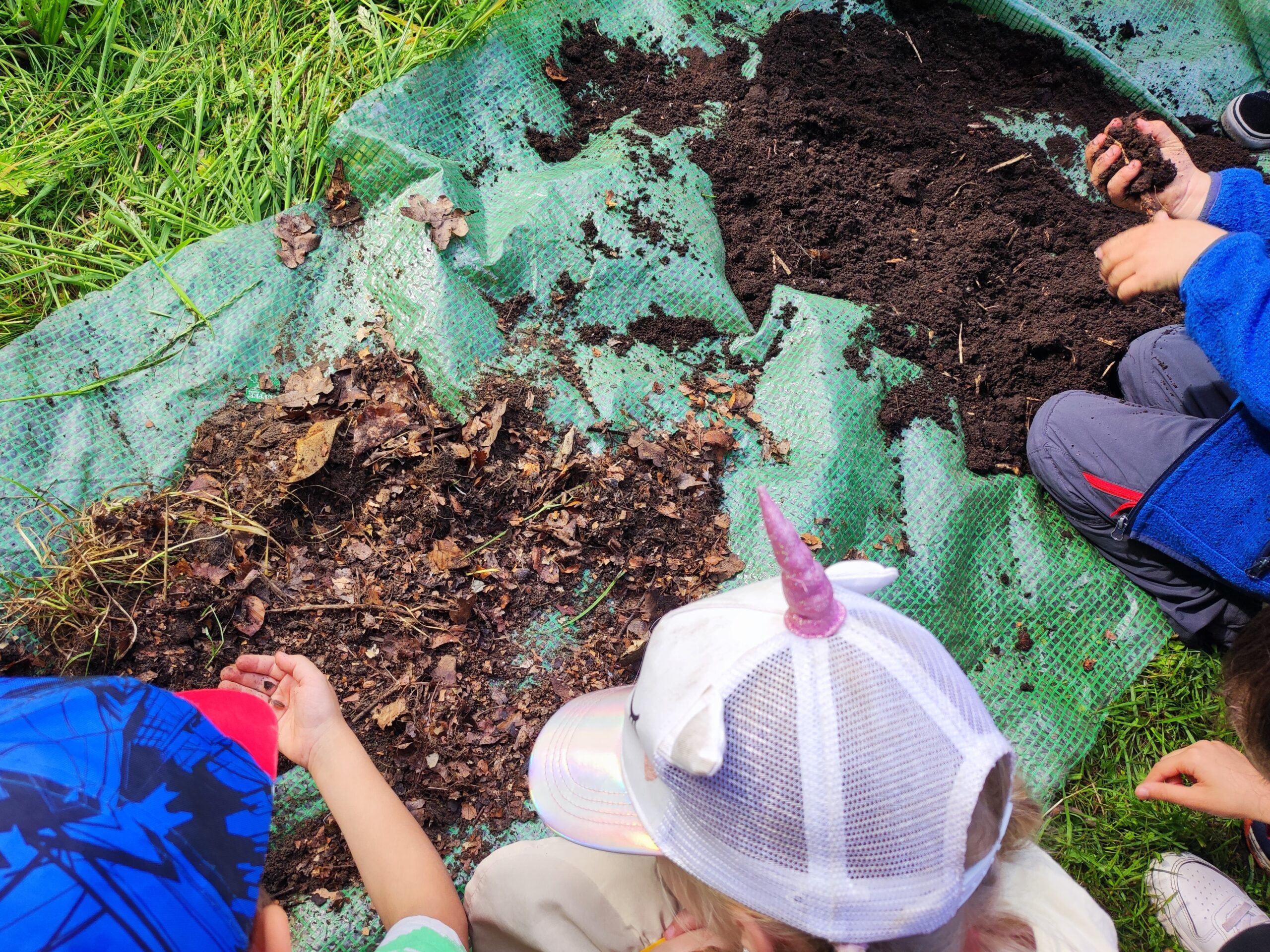 Im Garten Gehts Rund Kinder sind über einen Haufen feinen und einen Haufen groben Kompost gebeugt und untersuchen ihn. Ein Kind hat eine Assel in der Hand, ein anderes die feine Erde