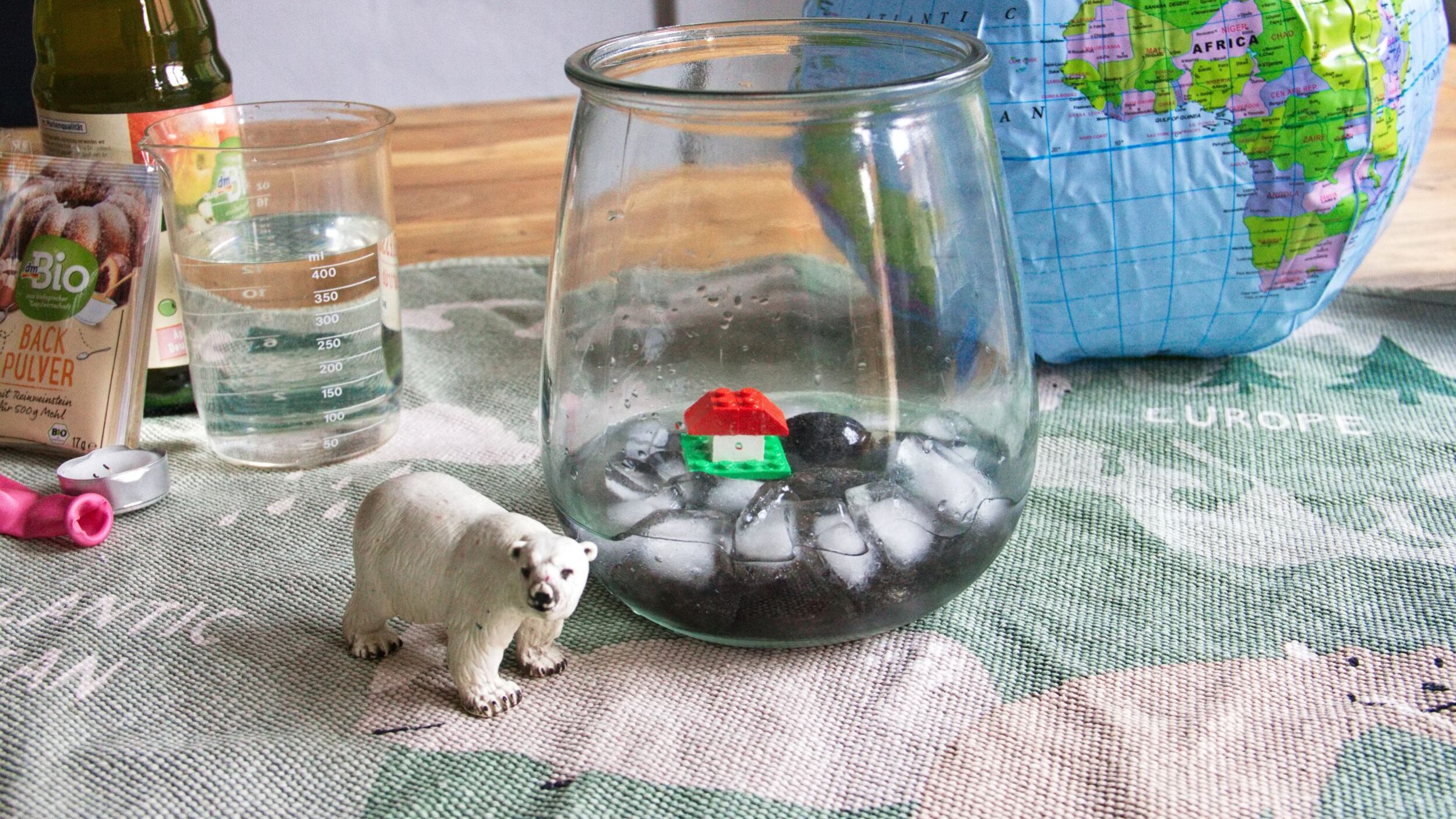 Auf einem Weltkarten-Teppich liegt eine aufblasbare Weltkugel, davor ein Weck-Glas mit Einswürfeln und einem kleinen Lego Haus auf einer Insel. Neben dem Glas steht ein Schleich-Eisbär. Im Hintergrund sieht man einen Messbecher, eine Essig-Flasche, eine Backpulvertüte und einen Luftballon