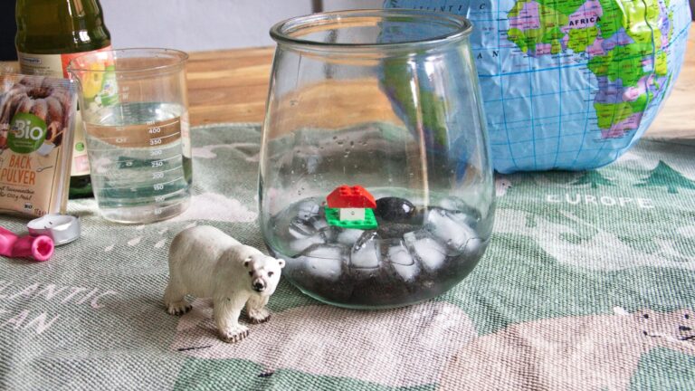 Auf einem Weltkarten-Teppich liegt eine aufblasbare Weltkugel, davor ein Weck-Glas mit Einswürfeln und einem kleinen Lego Haus auf einer Insel. Neben dem Glas steht ein Schleich-Eisbär. Im Hintergrund sieht man einen Messbecher, eine Essig-Flasche, eine Backpulvertüte und einen Luftballon