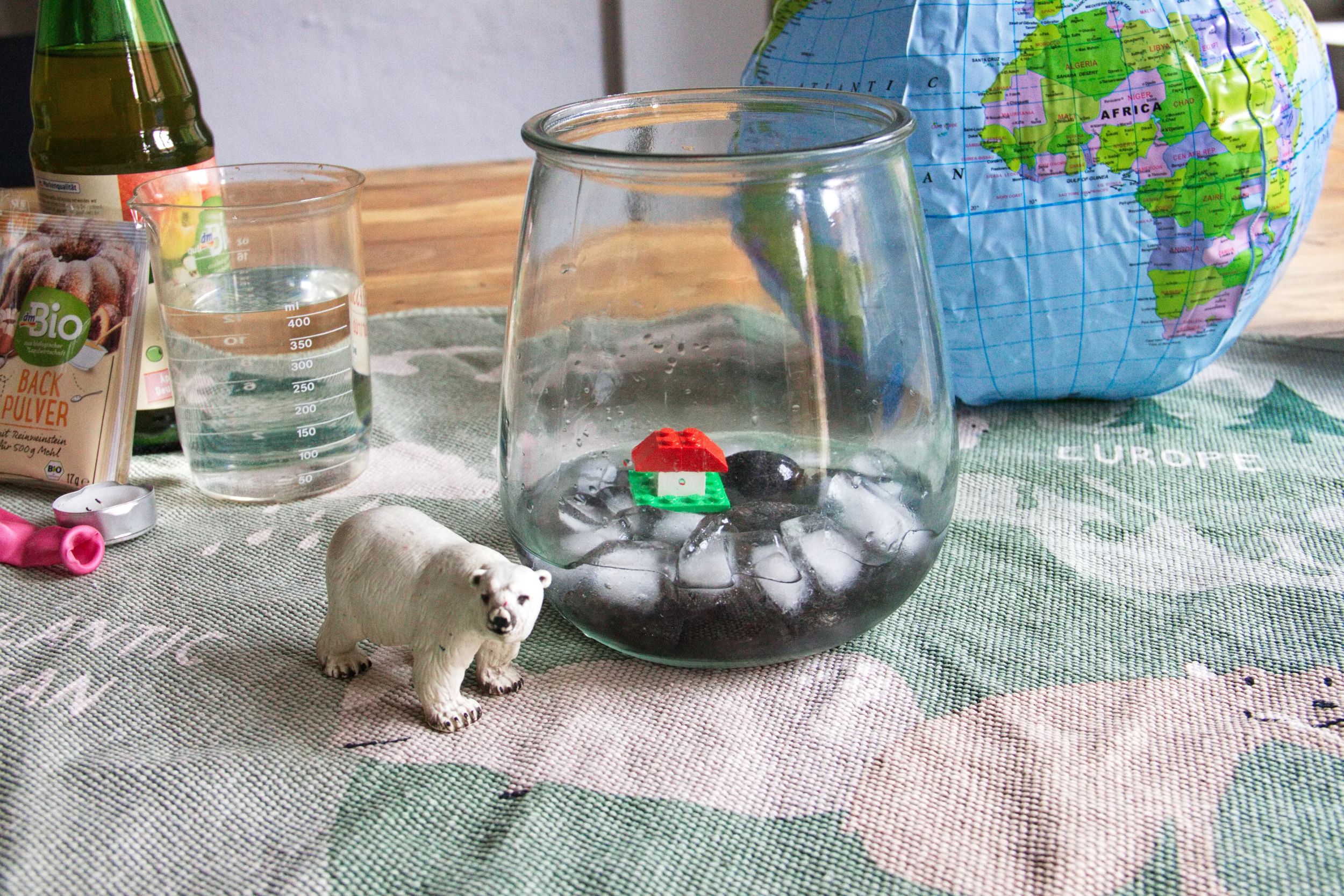 Klima Labor Auf einem Weltkarten-Teppich liegt eine aufblasbare Weltkugel, davor ein Weck-Glas mit Einswürfeln und einem kleinen Lego Haus auf einer Insel. Neben dem Glas steht ein Schleich-Eisbär. Im Hintergrund sieht man einen Messbecher, eine Essig-Flasche, eine Backpulvertüte und einen Luftballon